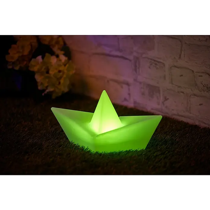 BAUHAUS Solar-Dekoleuchte Schiff - Weiß, IP65 - Leuchtendes Papierschiff auf Gras, mit sanftem grünem Licht, neben Blumen und einer Ziegelwand für stimmungsvolle Dekoration.