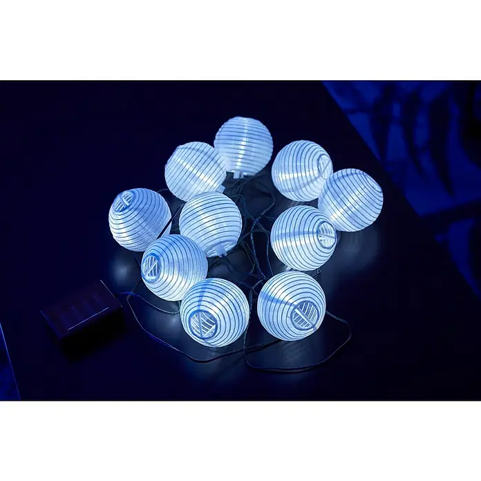 BAUHAUS Solar-Lichterkette Minilampions - Länge: 3,8 m, LED, Weiß - Nahaufnahme einer Lichterkette mit Solarpanel, bestehend aus zehn kugelförmigen, weißen Papierlaternen auf einem dunklen Tisch.