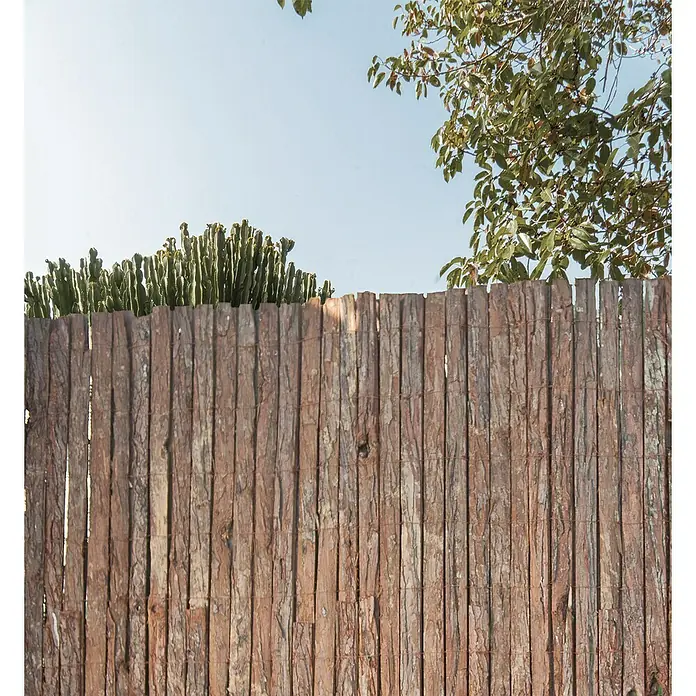 Faura Malla de ocultación corteza natural - 3 x 1 m, Marrón - Primer plano de una valla decorativa de madera con plantas y árboles al fondo bajo un cielo azul despejado.