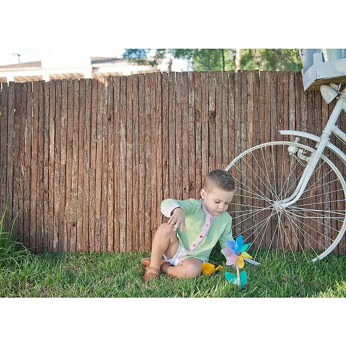 Faura Malla de ocultación corteza natural - 3 x 1,5 m, Marrón - Un niño pequeño está sentado en el césped jugando con un molinete de colores junto a una bicicleta vieja con canasta.