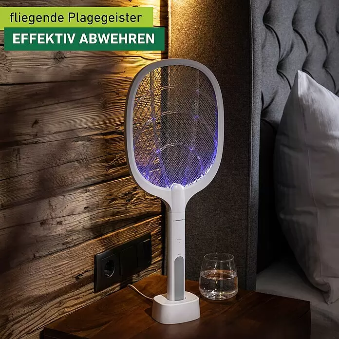 Windhager Elektrische Fliegenklatsche 2in 1 - L x B: 54 x 22 cm, Weiß - Weiße elektrische Fliegenklatsche mit blauem Licht auf Ladestation, auf einem Holznachttisch neben einem Bett.