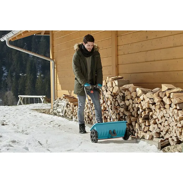Gardena Streuwagen L - Streubreite: 45 cm, 12,5 l - Ein Mann schiebt Schnee mit einer blauen Gardena Schneefräse vor einem Holzhaus. Praktisch und einfach für den Winterdienst.