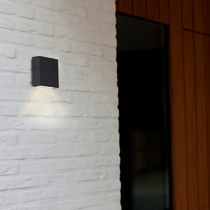 Lutec Aplique exterior LED Gemini XF - 9,5 W, 3,8 x 11 x 11 cm, Gris oscuro, IP54 - Lámpara de pared cúbica negra montada en una pared de ladrillo blanco, que emite un brillo cálido hacia abajo, un accesorio de iluminación elegante y moderno.