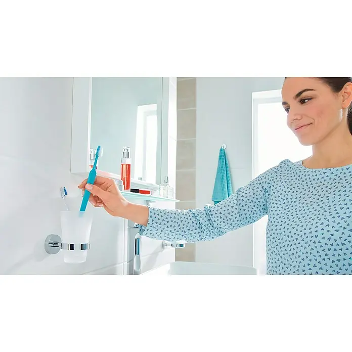 Tesa Smooz Portacepillos - Con soporte, Metal, Cromo satinado - Una mujer coge un cepillo de dientes de un portacepillos. El portacepillos está montado en la pared.