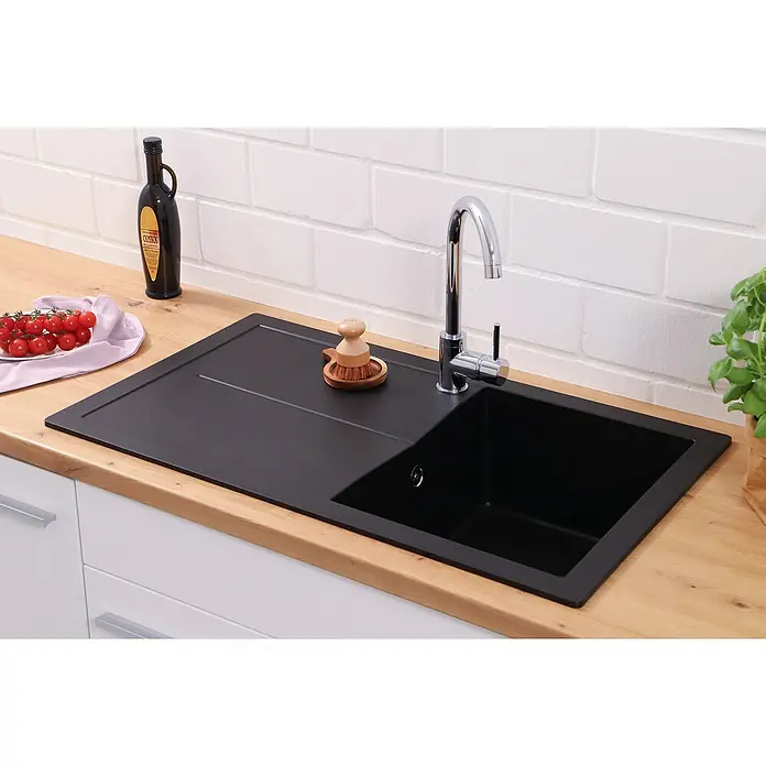 Respekta Einbauspüle Cleveland - 86 x 50 cm, Schwarz - Nahaufnahme einer schwarzen Spüle mit Abtropffläche, Armatur, Bürste, Tomaten und einer Flasche Öl auf einer hellen Holzoberfläche vor einer Ziegelwand.