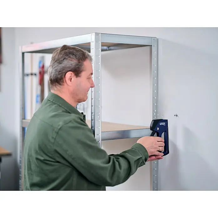 Rapid Montagezange XP30 - Passend für: Für Schrauben M4, M5 und M6 in Hohlwanddübeln - Ein Mann befestigt ein Regal mit einem blauen Rapid-Tacker an einer Wand. Das Regal ist aus Metall und hat mehrere Ebenen.
