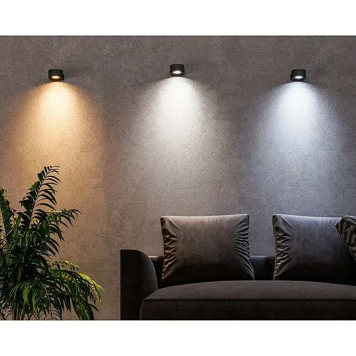 Lavida Akku-Wandleuchte Novara - L x B x H: 9,25 x 7,6 x 4,5 cm, 2 W, Schwarz, Mehrfarbig, Rund - Drei Wandleuchten mit unterschiedlichen Farbtemperaturen beleuchten eine graue Wand über einem dunklen Sofa und einer Pflanze.