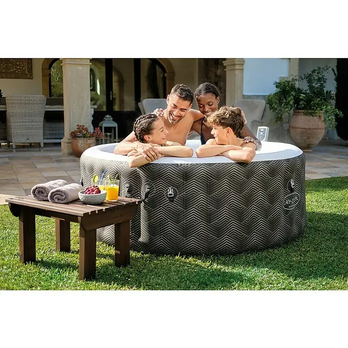 Bestway Lay-Z-Spa Mobil-Whirlpool Barbuda AirJet - Ø x H: 180 x 66 cm, Grau, 669 l - Das Bild zeigt einen Lay-Z-Spa Whirlpool mit vier Personen darin, Getränken und Handtüchern auf einem kleinen Tisch daneben.