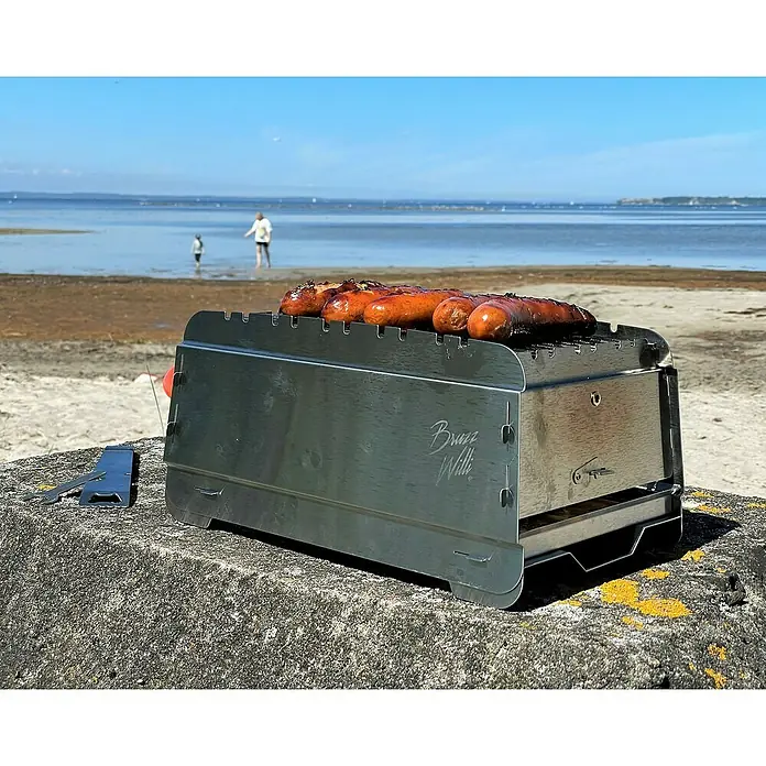 Bruzz Willi Gasgrill BW 1.1 - Wander Edition - Grillfläche (B x T): 39 x 19,5 cm, 3,5 kW, Anzahl Brenner: 1 Stk. - Ein tragbarer Grill mit Würstchen auf einem Stein, am Strand. Ideal für unterwegs, um leckere Würstchen zu grillen.