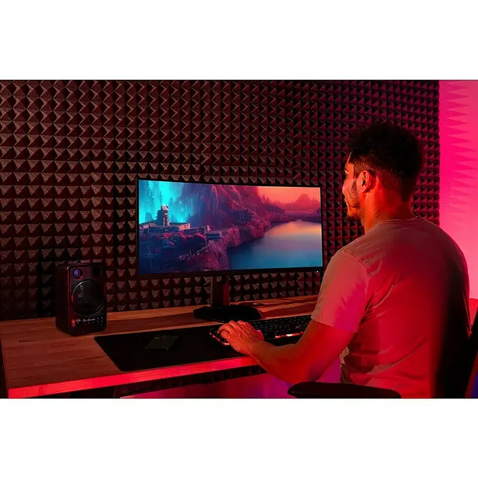 Noppenschaum Akupur - Pyramide, 100 x 50 x 4 cm, PU-Schaum - Ein Mann sitzt vor einem breiten Monitor und spielt ein Videospiel, umgeben von akustischen Paneelen und einer hellen Lautsprecherbox auf dem Tisch.