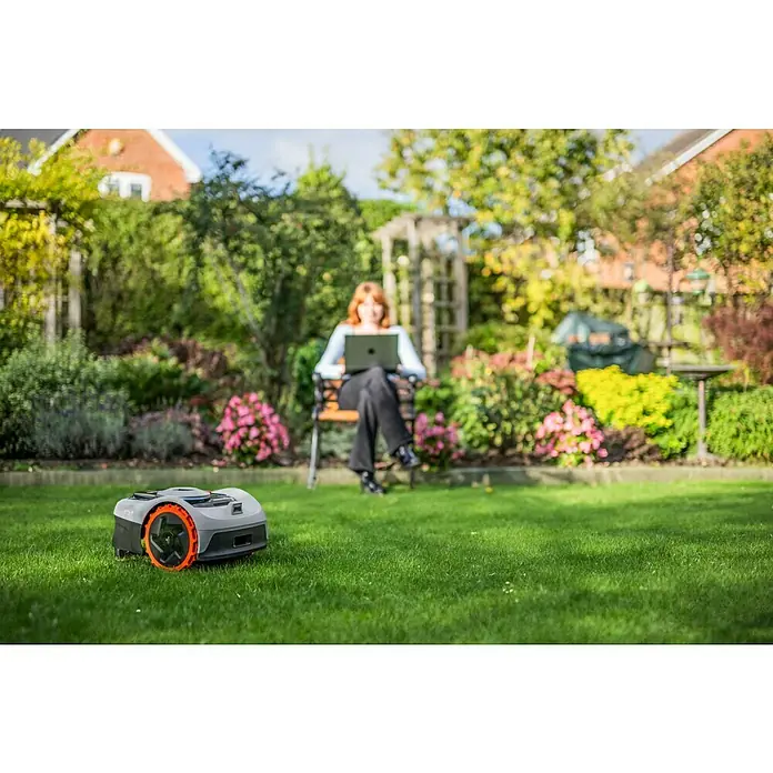 Segway Mähroboter Navimow i105E - 500 m², Schnittbreite: 18 cm, Satellitengestützte Navigation, RTK - Roboter-Rasenmäher mäht den Rasen, während eine Frau im Hintergrund mit ihrem Laptop arbeitet.