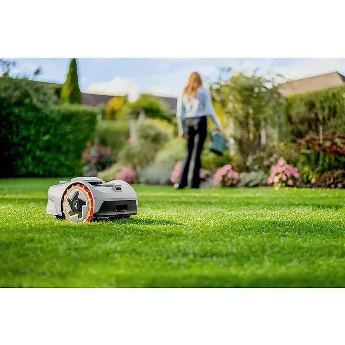 Segway Mähroboter Navimow i105E - 500 m², Schnittbreite: 18 cm, Satellitengestützte Navigation, RTK - Ein Rasenmähroboter steht auf einem grünen Rasen, im Hintergrund eine Frau mit Gießkanne, Büsche und ein Haus.