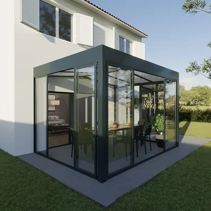 Wismar-NFI Wintergarten - 400 x 300 cm, Satteldach, Polycarbonat, Anthrazitgrau, Milchig Weiß - Bild eines Wintergartens aus Glasanbau, der sich an einem Wohnhaus befindet und einen Essbereich mit Tisch und Stühlen bietet.
