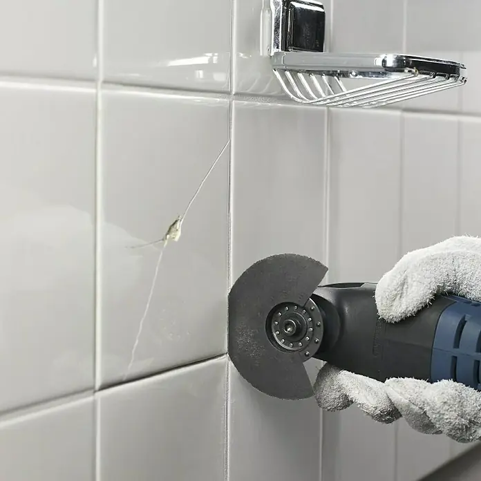 Bosch Zubehör-Set Fliesen - 3 Stk., 3 -tlg. - Eine Hand mit Schutzhandschuh entfernt mit einer oszillierenden Multitool-Säge eine beschädigte Fliese an einer Badezimmerwand.