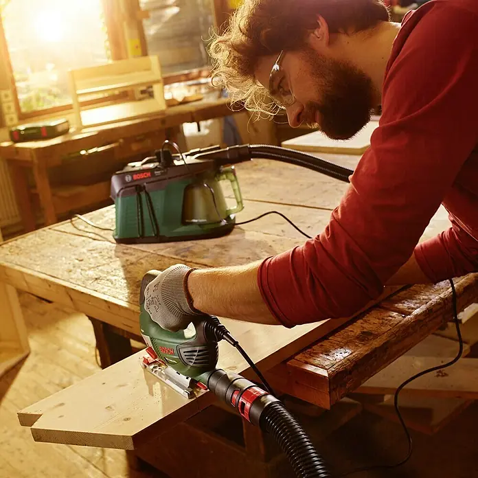 Bosch Elektro-Staubsauger EasyVac 3 - 700 W, Behältervolumen: 2,1 l - Ein Mann benutzt eine Bosch Stichsäge mit Staubabsaugung in einer Werkstatt, um ein Holzbrett zu bearbeiten.