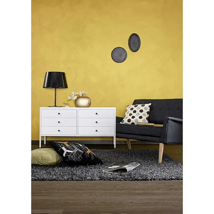 SCHÖNER WOHNEN-Farbe Trendstruktur Farbeffektputz Metall-Optik Effektfarbe - Gold, Glänzend, 2,5 l - Elegantes Wohnzimmer mit einem weißen Sideboard, einer schwarzen Lampe und einem grauen Sessel auf einem flauschigen Teppich. Die gelbe Wand verleiht Wärme.