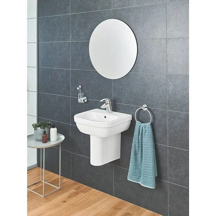 Grohe Eurosmart Grifo de lavabo - Cromo, Brillante, Con tirante, Arranque en agua fría - Una moderna escena de baño con un lavabo blanco, un espejo redondo y azulejos grises.