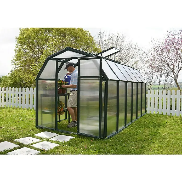 Palram – Canopia Invernadero Eco Grow - Medidas ext. (An x Pr): 204 x 383 cm, 6 mm, Verde - Hombre regando plantas en un invernadero con marcos verdes y paredes transparentes, rodeado de un jardín.