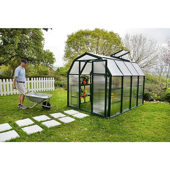 Palram – Canopia Invernadero Eco Grow - Medidas ext. (An x Pr): 204 x 239 cm, 6 mm, Verde - Hombre con una carretilla frente a un invernadero en el jardín. El invernadero es de vidrio y metal y alberga plantas.