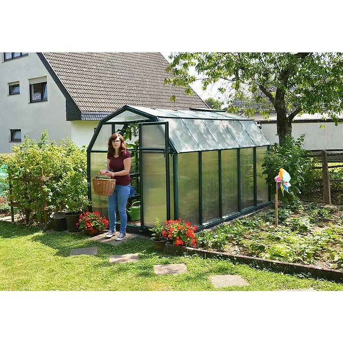 Palram – Canopia Invernadero Eco Grow - Medidas ext. (An x Pr): 204 x 321 cm, 6 mm, Verde - Primer plano de un invernadero en el jardín, con una mujer sosteniendo una cesta, rodeada de plantas y flores para jardinería relajada.