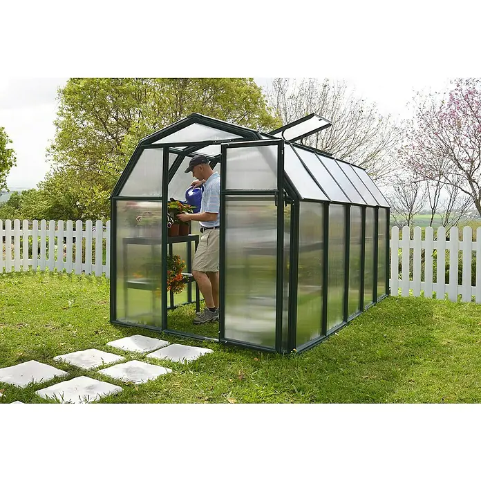 Palram – Canopia Invernadero Eco Grow - Medidas ext. (An x Pr): 204 x 321 cm, 6 mm, Verde - Un hombre riega las plantas en un invernadero. El invernadero está en el jardín con una valla blanca al fondo.