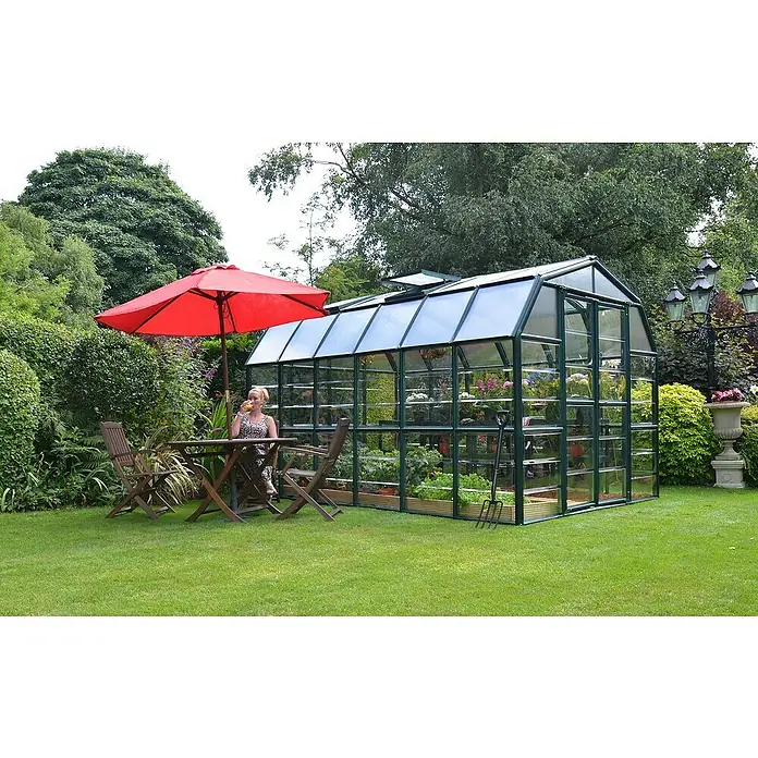 Palram – Canopia Invernadero Grand Gardener - Medidas ext. (An x Pr): 264,7 x 388,6 cm, Material de la pared lateral: Plástico, 0,8 mm, Verde - La imagen muestra un invernadero de vidrio y metal en un jardín, con una mujer sentada a una mesa bajo una sombrilla roja.