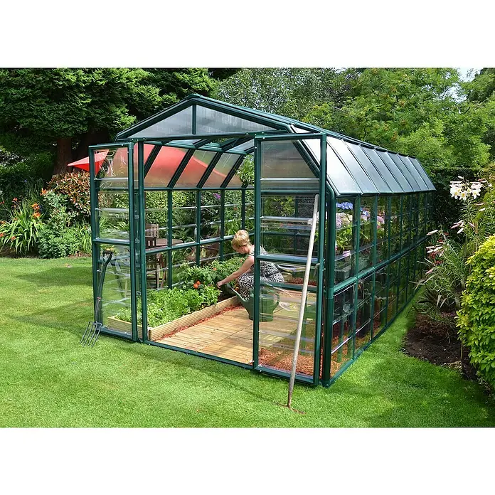Palram – Canopia Invernadero Grand Gardener - Medidas ext. (An x Pr): 264,7 x 513 cm, Material de la pared lateral: Plástico, 0,8 mm, Verde - Invernadero con borde verde en un jardín, una mujer riega las plantas en el interior, destacando su estructura y funcionalidad.