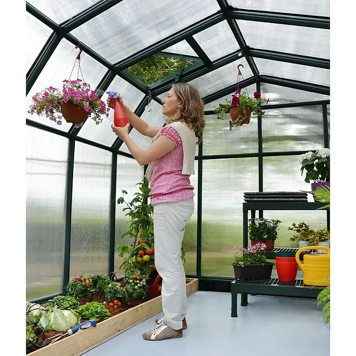 Palram – Canopia Invernadero Hobby Gardener - Medidas ext. (An x Pr): 266 x 266 cm, 6 mm, Verde - Una mujer rocía flores en un invernadero, rodeada de exuberante vegetación y diversas plantas que florecen en macetas y parterres elevados.