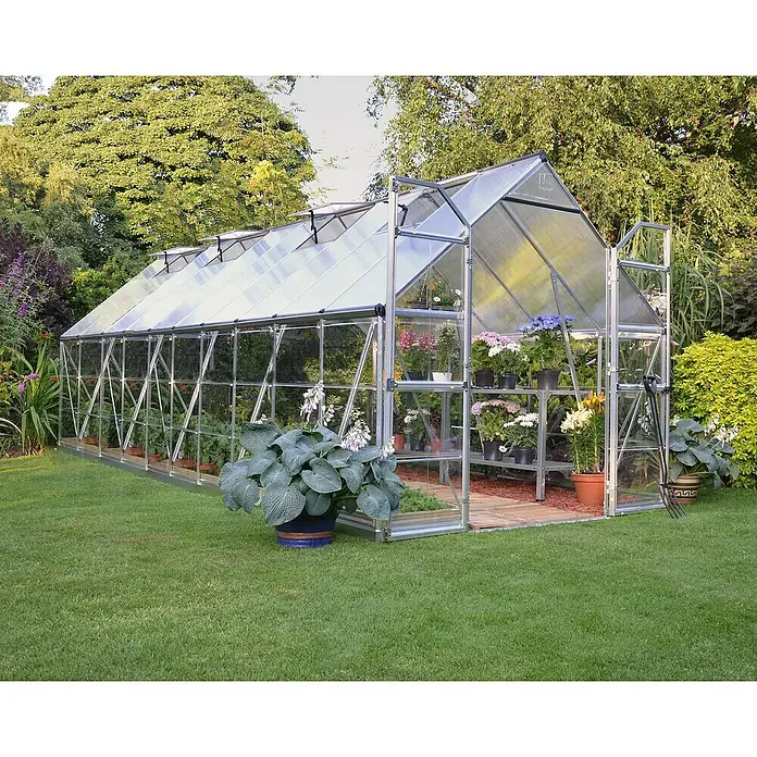Palram – Canopia Invernadero Balance - Medidas ext. (An x Pr): 244 x 607 cm, 0,7 mm, Plateado - Un espacioso invernadero con plantas, macetas y herramientas de jardinería rodeado de exuberante vegetación. ¡Ideal para amantes del jardín!
