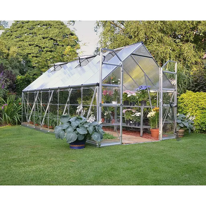 Palram – Canopia Invernadero Balance - Medidas ext. (An x Pr): 244 x 487 cm, 0,7 mm, Plateado - Un espacioso invernadero de aluminio con una puerta abierta, rodeado de exuberante vegetación en el jardín, proporciona una protección ideal para las plantas.