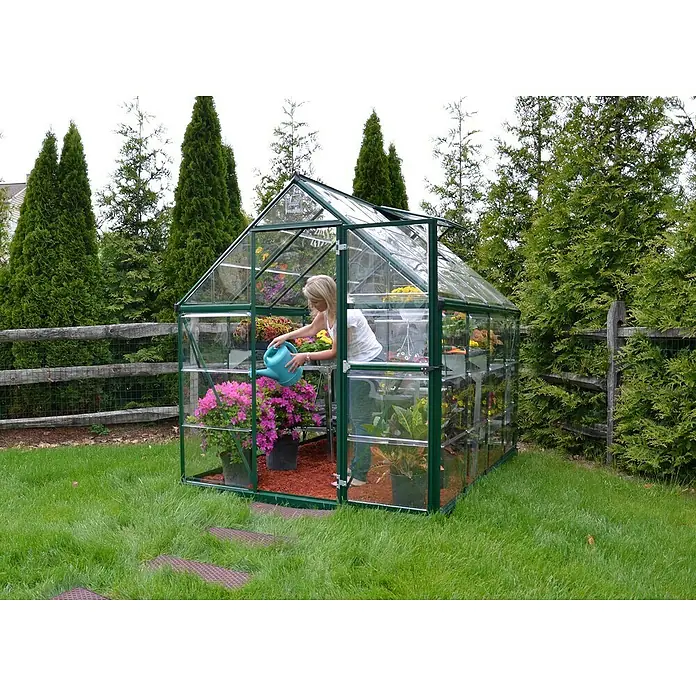 Palram – Canopia Gewächshaus Harmony - Außenmaß inkl. Dachüberstand (B x T): 185 x 247 cm, Grün, Grundfläche: 4,6 m² - Frau gießt Pflanzen in einem geräumigen Gewächshaus, das mit Grünpflanzen und bunten Blumen gefüllt ist.