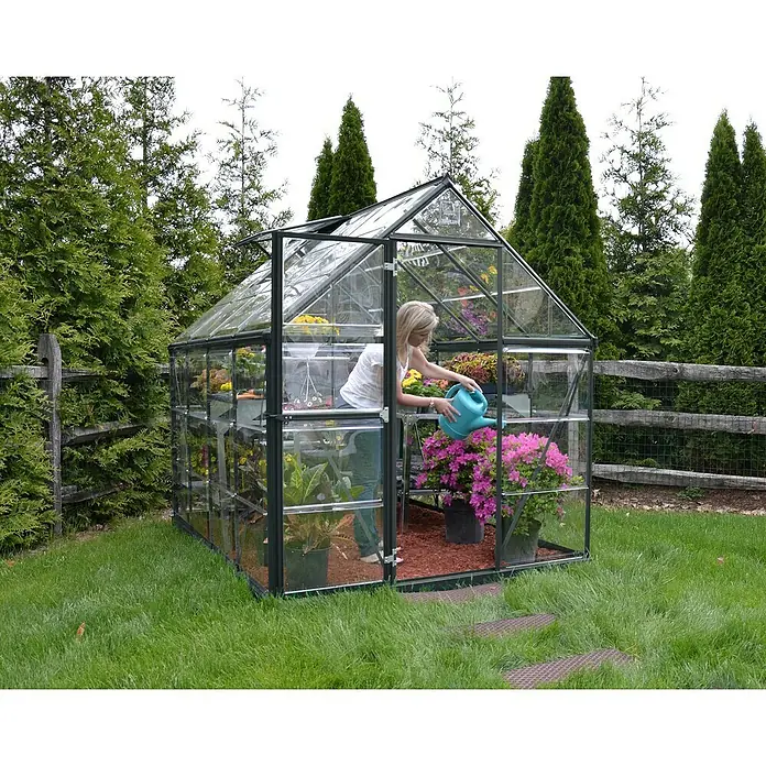 Palram – Canopia Invernadero Harmony - Medidas ext. (An x Pr): 185 x 247 cm, Gris, Área: 4,6 m² - Mujer regando plantas en un invernadero. El invernadero es de vidrio y metal y está situado en un jardín.