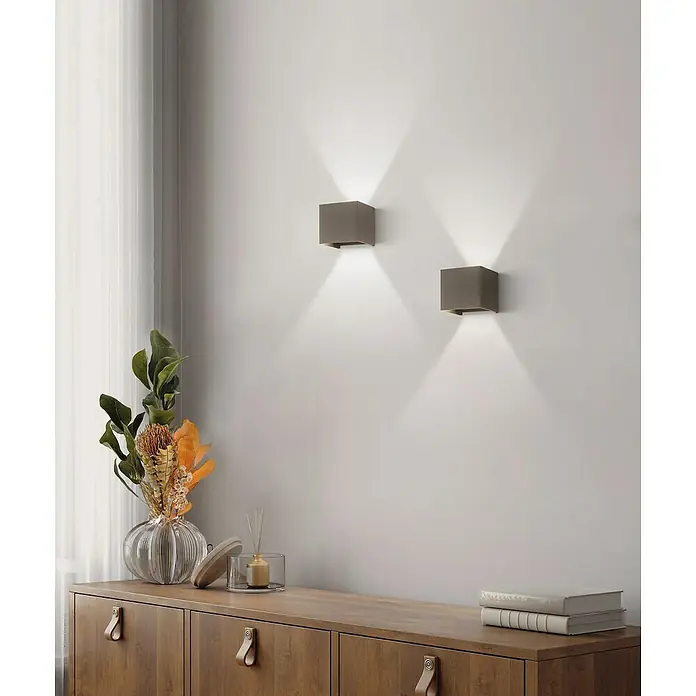 Fischer & Honsel LED-Außenwandleuchte Wall - 6 W, 10 x 11 x 10 cm, Schwarz, IP44 - Wandbild mit zwei quadratischen Lampen, die ein angenehmes Licht abstrahlen, und einer dekorativen Kommode mit Blumen und Büchern.
