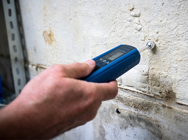 Wand trocknen nach Wasserschaden: Hand hält Feuchtigkeitsmessgerät an eine Wand