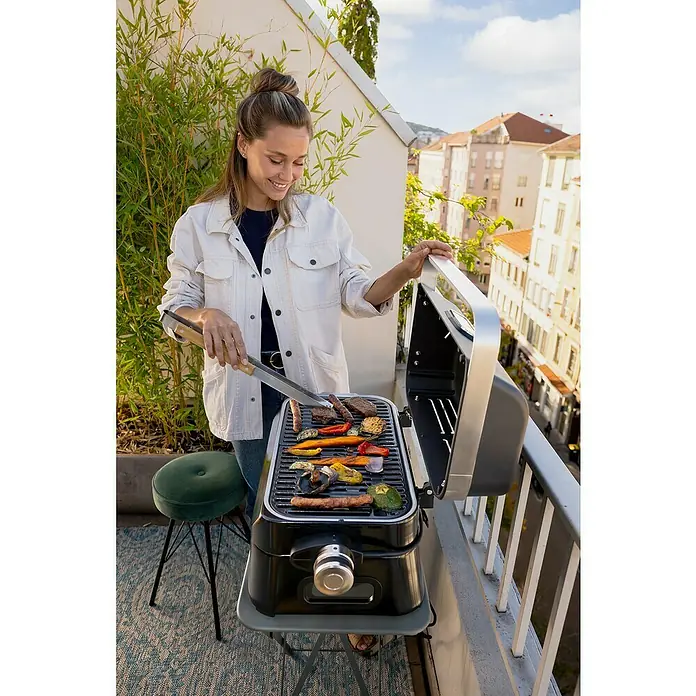 Campingaz Barbacoa eléctrica Attitude 2GO - Superficie de la parrilla (An x Pr): 48 x 26 cm, 2.200 W - Una mujer asa filetes y verduras en una parrilla eléctrica de mesa en un balcón con vistas a la ciudad.