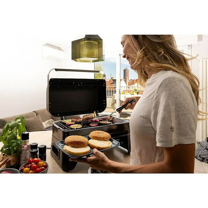 Campingaz Barbacoa eléctrica Attitude 2GO - Superficie de la parrilla (An x Pr): 48 x 26 cm, 2.200 W - Mujer preparando hamburguesas con una parrilla portátil llena de verduras y carne, con dos panes de hamburguesa cerca.