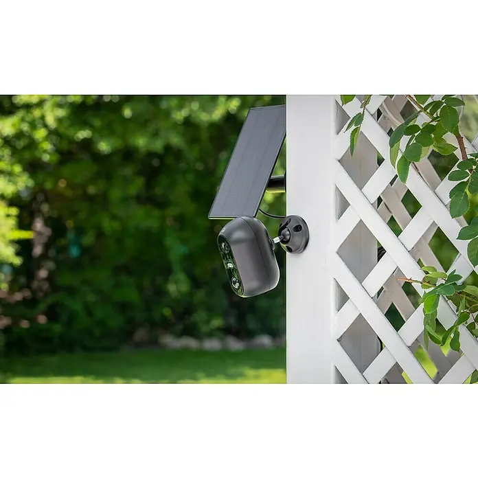 Garza Smart Home Cámara de vigilancia solar Dalia 200 - Negro, Ángulo de detección: 140 ° - Una cámara de seguridad inalámbrica con panel solar está montada en un enrejado blanco, ofreciendo vigilancia exterior.