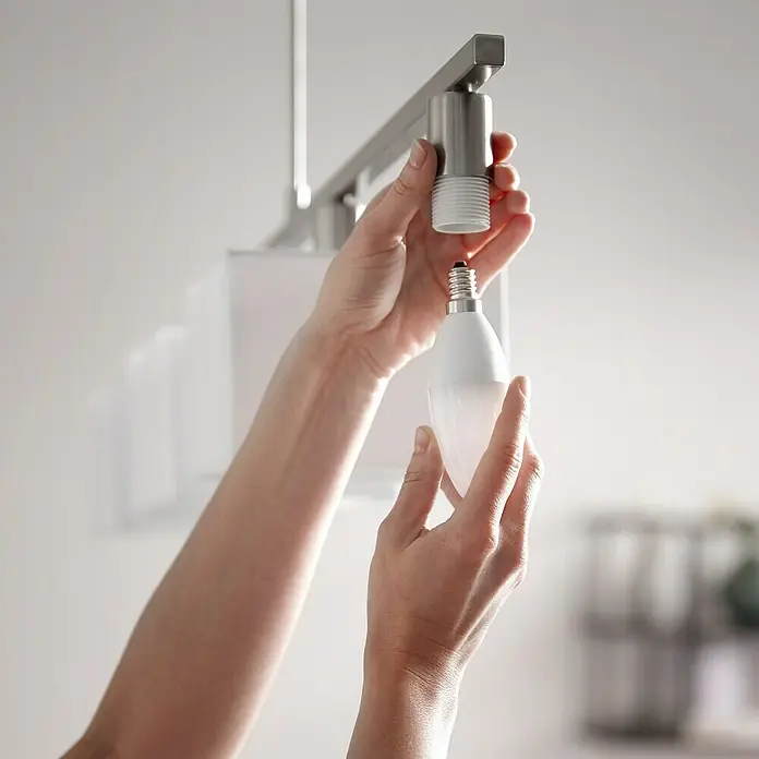 Philips Hue Bombilla LED RGB - E14, Intensidad regulable, RGBW, 470 lm, 6,5 W - Una mano enrosca una bombilla LED blanca en un portalámparas plateado sujeto a un riel metálico. Iluminación interior moderna.