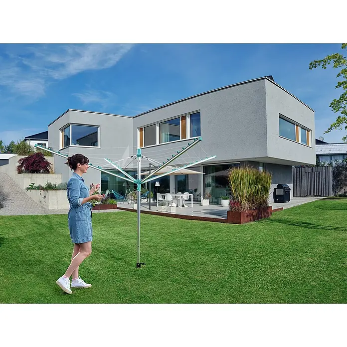 Leifheit Wäschespinne Linomatic 500 Easy Wäscheschirm - Geeignet für Anzahl Waschmaschinenfüllungen: 5 Stk., Länge Wäscheleine: 50 m, Durchmesser offen: 3,28 m, Grau/Metallic, Mint - Eine Frau bedient einen grünen Wäscheständer im Garten eines modernen Hauses mit strahlend blauem Himmel.