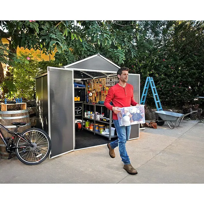 Palram – Canopia Gartenhaus Rubicon - Außenmaß inkl. Dachüberstand (B x T): 1,85 x 3,78 m, Kunststoff, Grau - Ein Mann trägt eine Kiste mit Gegenständen aus einem geräumigen Gartenschuppen mit Regalen und Werkzeugen.
