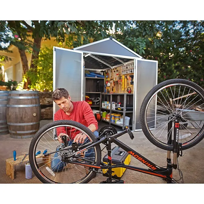 Palram – Canopia Gartenhaus Rubicon - Außenmaß inkl. Dachüberstand (B x T): 1,85 x 3,78 m, Kunststoff, Grau - Ein Mann repariert ein Fahrrad vor einem Werkzeugschuppen. Werkzeuge und Zubehör sind ordentlich im Schuppen verstaut, was die Organisation erleichtert.