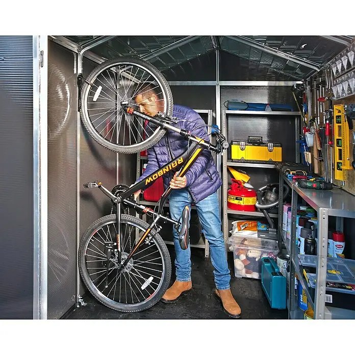 Palram – Canopia Gartenhaus Rubicon - Außenmaß inkl. Dachüberstand (B x T): 1,85 x 3,78 m, Kunststoff, Grau - Ein Mann hängt ein Rainbow Fahrrad in einem Schuppen auf, der voll mit Werkzeugen und Gegenständen ist.