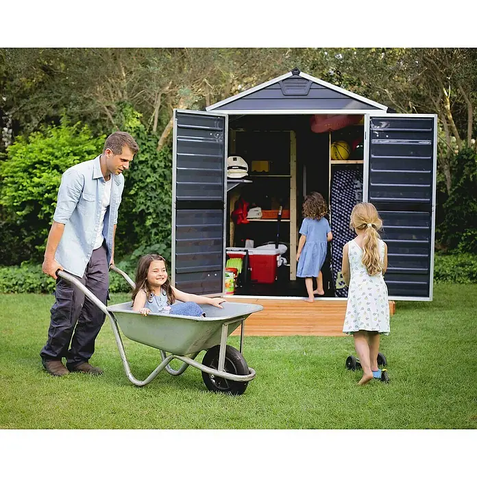 Palram – Canopia Caseta de jardín Skylight - Medidas ext. (An x Pr): 185 x 229 cm, Plástico, Antracita - Un padre empuja a su hija en una carretilla en el jardín delante de un nuevo cobertizo. Dos niños juegan en el jardín.