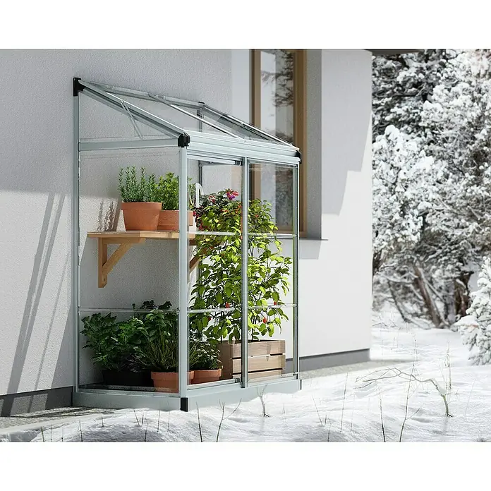 Palram – Canopia Invernadero Lean To - Medidas ext. (An x Pr): 125 x 63,5 cm, Material de la pared lateral: Plástico, 0,7 mm, Plateado - Primer plano de un invernadero adosado con plantas en un estante contra un fondo nevado.