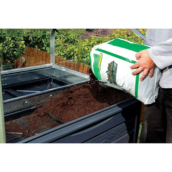 Palram – Canopia Invernadero Plant Inn - Medidas ext. (An x Pr): 118 x 118 cm, Material de la pared lateral: Plástico, 0,7 mm, Antracita - Un hombre llenando un huerto elevado en un invernadero con tierra de un saco. El huerto elevado es de madera oscura.