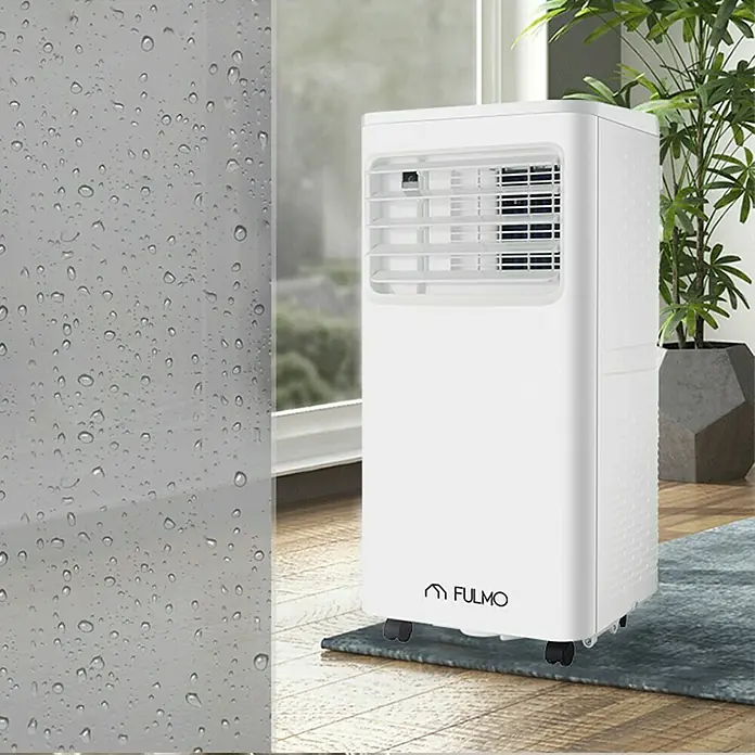 Fulmo Aire acondicionado portátil - 3.500 W, Tamaño de la estancia: 30 m², Blanco - Un aire acondicionado Fulmo blanco y portátil sobre ruedas, colocado sobre una alfombra en una habitación luminosa con una planta y una ventana.