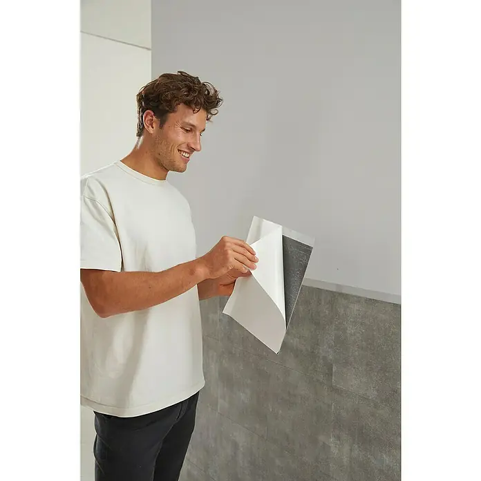 D-c-fix Fliesensticker - 6 Stk., 33,5 x 33,5 cm, Solid Concrete - Ein Mann, der eine selbstklebende Wandfliese anbringt, zeigt die einfache Installation und das moderne Design der grauen Fliese.