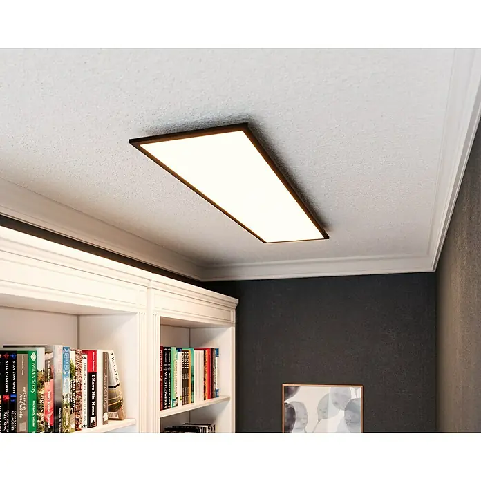 Tween Light LED-Panel CCT-RC-DIM - 32,5 W, L x B x H: 120 x 30 x 5 cm - Eine rechteckige Deckenleuchte mit schlankem, dunklem Rahmen erhellt einen Raum mit Bücherregal und Kunstwerk.