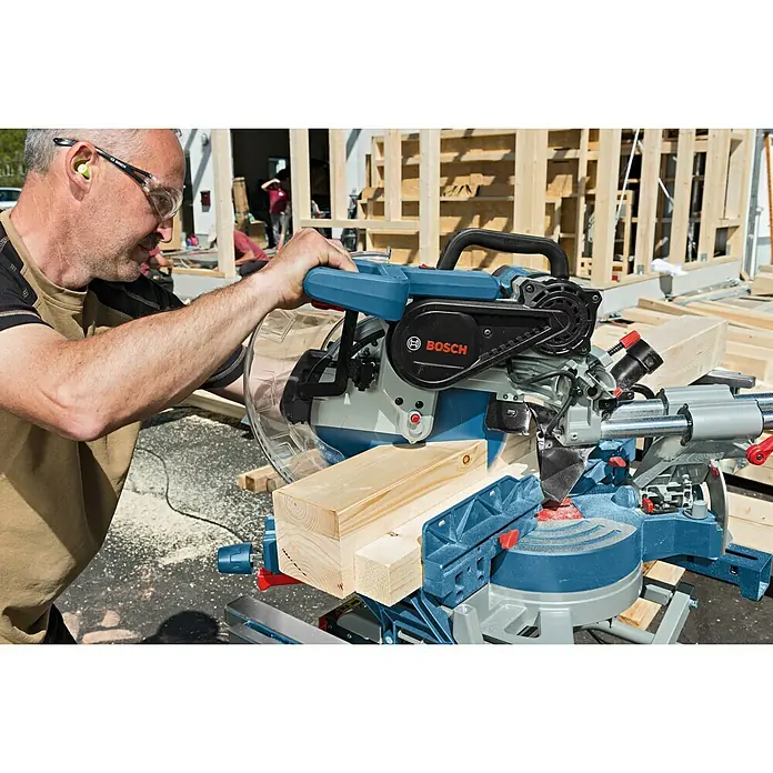 Bosch Professional Paneelsäge GCM 12 SDE - 1.800 W, 3.100 U/min - 4.000 U/min, Durchmesser Sägeblatt: 305 mm, Schnitttiefe: 85 mm bei 90 °/90 ° - Ein Mann bedient eine Bosch Kapp- und Gehrungssäge, um Holz zu schneiden. Das Gerät ist blau und grau und scheint in gutem Zustand zu sein.