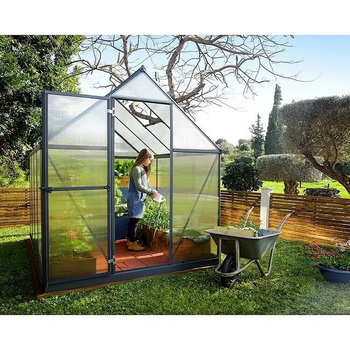 Palram – Canopia Invernadero Mythos - Medidas ext. (An x Pr): 185 x 247 cm, 4 mm, Gris - Mujer riega plantas en un invernadero con estructura de acero y paredes transparentes. En el jardín, hay una carretilla llena de herramientas de jardinería.